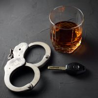 concept of criminal liability for driving while intoxicated. Bakal with cognac, handcuffs, car key on a dark background.